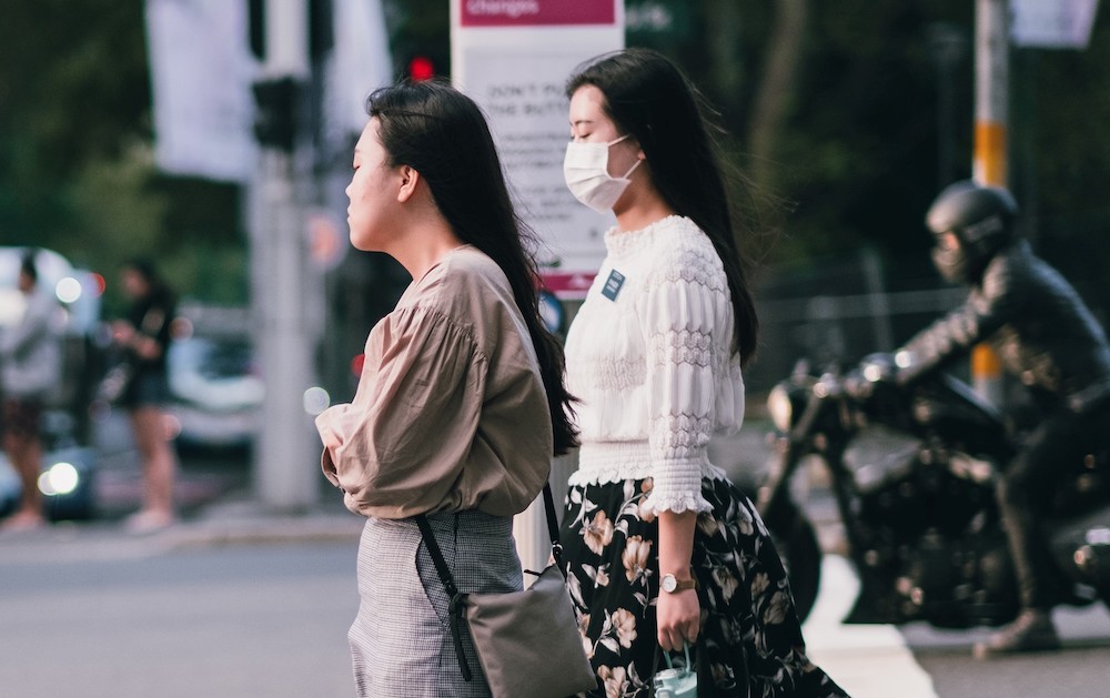 two women in masks wait to cross the street