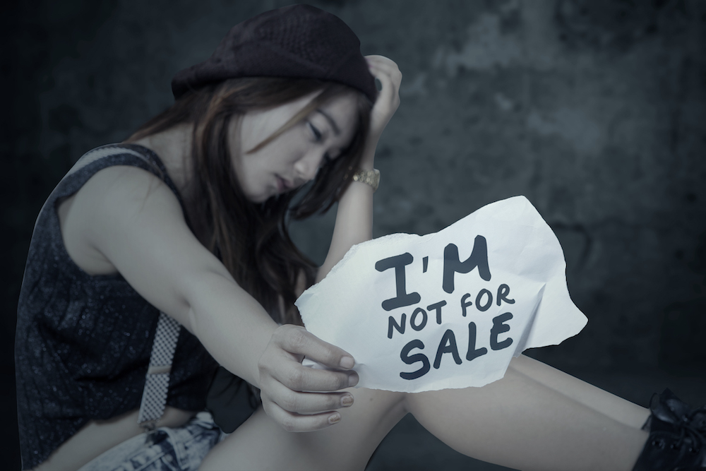 a girl sits with her head bowed and hand on top of her head, holding a sign saying "I'm not for sale"