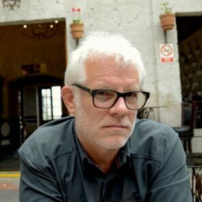 A man with white hair and black glasses in front of a doorway